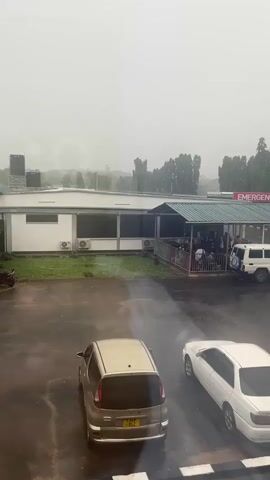 A high-angle, long shot shows the emergency room entrance of a hospital on a rainy day. Several vehicles are parked in the lot in front of the entrance, including a tan hatchback, a white sedan, a red hatchback next to a motorcycle, and a white SUV. A row of tall, thin trees lines the back of the parking lot. People are gathered underneath the covered entrance of the emergency room.