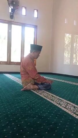 A man wearing a red patterned shirt and a black cap sits cross-legged on a green carpet in what appears to be a mosque. He is facing away from the camera, towards a large window. The carpet has a decorative border. The room has a high ceiling with rectangular windows. There is a fan mounted on the wall.