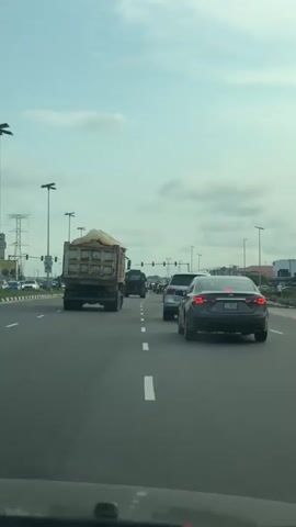 A rear view of traffic on a highway. A large dump truck loaded with sand is in the left lane. A dark-colored sedan is in the right lane behind the dump truck. Ahead of the sedan, a silver SUV is visible. Other vehicles and streetlights line the highway, extending into the distance. The sky is overcast.