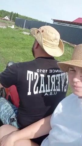 Here's a description of the video:

The video shows a man and a woman riding a small motorcycle with a trailer. The man is driving and wearing a black t-shirt with the word "EVERLAST" printed backwards and a light-colored bucket hat. He also has a beard. He is wearing blue camouflage pants. The woman is riding behind him, wearing a white t-shirt and a straw hat. She is filming the video.

The trailer is full of birch logs and a chainsaw. They are driving on a dirt road through a rural area. Houses and power lines are visible in the background. The day appears sunny.