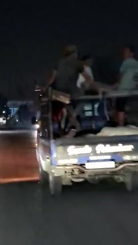 A pickup truck drives down a road at night. Three people are sitting in the bed of the truck, facing forward. The truck has writing on the tailgate that is difficult to read. The road is paved and there are streetlights visible in the background. There is also a bright light from an oncoming vehicle.