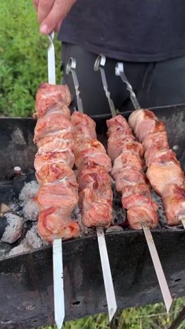 A person is grilling kebabs over a charcoal grill. There are four metal skewers with chunks of meat on them, arranged in a line on the grill. The meat is pink and appears to be cooking. The grill is black and rectangular, and the coals underneath are red and glowing. The background is out of focus but appears to be an outdoor setting with greenery. The person is wearing a black t-shirt and dark pants, and their hands are visible as they adjust the skewers. The video is short and shows the kebabs rotating slightly on the grill.