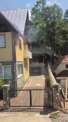 The video shows a view from a moving vehicle, likely a car or bus, looking out a window. The camera pans across a residential street, focusing on houses and vegetation. The first house visible is a two-story yellow building with a stone facade on the lower level. It has a gray roof, multiple windows, and a balcony. A black metal gate and a stone wall with a rose bush in front of it are also visible. The vehicle continues to move, and the view shifts to a larger, light-colored house with a red roof in the background. A blue swimming pool is visible in the yard of this second house, surrounded by a fence. Lush green trees and bushes are abundant throughout the scene, suggesting a summer day. The sky is blue with some clouds. There are no people or animals visible in the video. The quality of the video is somewhat obscured by what appears to be dirt or smudges on the window.