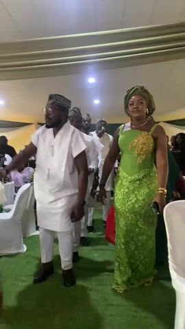 The video shows a man and woman walking together in a celebratory procession, possibly at a wedding or special event. The man is wearing a traditional white outfit with a cap, and the woman is in a green lace dress with a matching head wrap. They are surrounded by other people, also in traditional attire, some of whom are dancing and smiling. A photographer is taking pictures of the couple. The setting appears to be an indoor event space with white chairs and decorations.