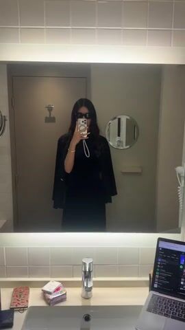 In a medium-shot video, a woman stands in front of a large, rectangular mirror in a bathroom, taking a selfie with her smartphone. The mirror is backlit and surrounded by white square tiles. The woman has long dark hair, and she is wearing sunglasses, a black dress, a white pearl necklace, a gold bracelet, and a black blazer draped over her shoulders. She is holding her smartphone up to her face with her right hand, and her left hand is resting on her hip. Behind her, a closed door is visible to the left, and a round mirror is on the right. On the counter below the mirror, there are various items, including a stack of cards, a silver container, and a laptop. The laptop screen displays various open windows. The bathroom has a neutral color scheme, and the lighting is bright.