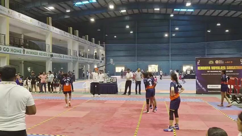 A group of young women are playing kabaddi indoors. The court is a red and blue mat with yellow lines. One player in an orange and blue uniform is running across the court. Several other players in similar uniforms are on the court, some facing her, some facing away. A crowd of spectators watches from bleachers. In the background, banners display "Kendriya Vidyalaya Sangathan" and "National Sports Meet."