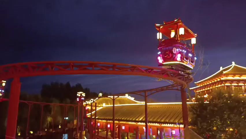 A red roller coaster with a train decorated with Chinese lanterns moves along a track at night. In the background, traditional Chinese buildings are illuminated with lights and red lanterns. Trees and foliage are also visible. The sky is dark blue, suggesting it is dusk or night.