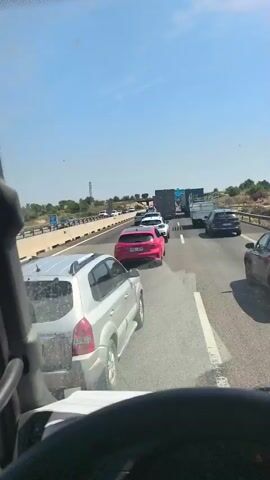 From inside the cab of a large vehicle, a congested highway is visible. Many cars, trucks, and vans are stopped or moving very slowly. There is a pink hatchback, a white hatchback, and a silver SUV directly in front of the camera. The sky is blue and mostly clear, and the vegetation alongside the road is sparse. The sun is out. The view is mostly static.