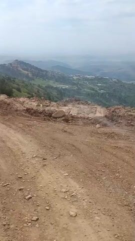 The video shows a dirt road with a vehicle's dashboard visible in the foreground. The road is on a hillside overlooking a valley. The valley is green and appears to be agricultural. The sky is overcast. The camera appears to be mounted inside a vehicle, possibly a truck or SUV, looking out the windshield. The vehicle is moving along the dirt road. The road itself appears to be rough and unpaved, with loose rocks and dirt. The terrain is mountainous, with hills and mountains visible in the distance. There are some trees and shrubs growing on the hillsides.