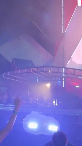The video shows an outdoor music festival at night. A DJ is performing on a stage under a large, abstract-looking canopy structure. Lights illuminate the stage and the crowd. The sky is dark, and trees are visible in the background. The camera pans slightly and focuses on the DJ and the stage. The video captures a lively atmosphere with people enjoying the music.