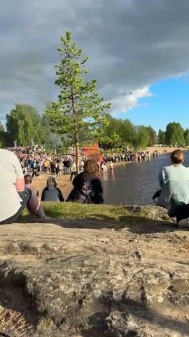 The video shows a large bonfire burning on a sandy shore next to a body of water. A crowd of people is gathered around the bonfire, some standing on the beach and others wading in the water. The sky is partly cloudy, and the sun is shining, casting a warm glow on the scene. The bonfire is large and emits a lot of smoke and flames, creating a dramatic effect. The people in the video appear to be enjoying the event, with some children seen playing in the water. The overall atmosphere seems festive and celebratory.
