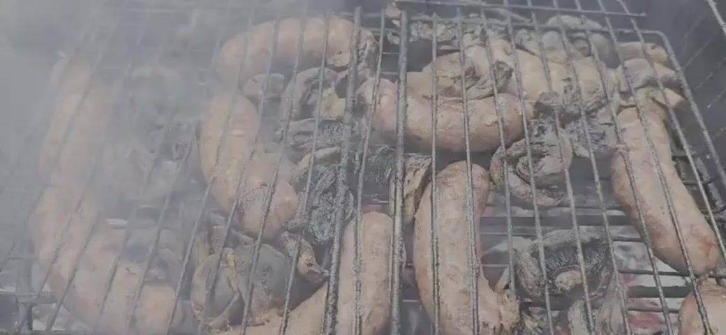 The video shows an extreme close-up, overhead shot of sausages and mushrooms being grilled on a metal rack. Smoke drifts across the scene, obscuring some of the food. The sausages are a light brown color, and some appear to be slightly charred. The mushrooms are darker and appear to be whole. The metal grill bars are visible throughout the shot, and the food is arranged in rows on the grill. The lighting suggests an outdoor environment, possibly a sunny day, though the overall tone is muted by the smoke. The camera is positioned directly above the grill, looking down at the cooking food.