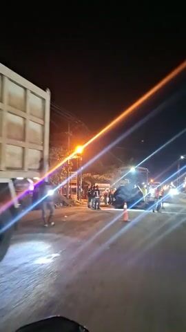 A night-time video shows a street scene with multiple light sources creating lens flares. Several people are visible, some wearing vests that suggest they are workers. A large piece of construction equipment, possibly a paver or a similar machine, is present on the road. Orange traffic cones are also visible. The ground appears wet or reflective, possibly from recent rain or the construction activity itself. The overall impression is of road work or maintenance being conducted at night.
