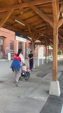 The video begins by showing a wooden roof structure above a train platform. A woman with dark hair, wearing a red shirt, blue jacket, and leopard-print leggings, pushes a stroller containing a baby. She is walking away from the camera, towards the right side of the frame. To her left, a man in a black t-shirt and dark pants stands with his back to the camera, facing a sign. The sign is blue and displays text in white, indicating "Kamienna Góra" at the top. Below this, two directional arrows point left and right, with text that appears to be station names or destinations, though it is not entirely clear. The building behind the sign is made of red brick and has graffiti on its walls. The platform itself is paved, and there are white poles supporting the wooden roof. The overall impression is a somewhat weathered but functional train station environment.