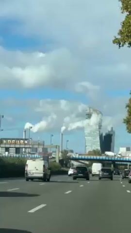The video shows a multi-lane highway with moderate traffic on a partially cloudy day. In the background, there are tall buildings and industrial smokestacks emitting white smoke. The shot appears to be taken from within a moving vehicle.