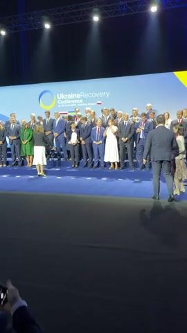 The video shows a group of people on a stage in front of a large screen with the text "UkraineRecovery Conference 25-26 June 2026 - Gdańsk, Poland". The screen also features a blue and yellow circular logo, representing the Ukrainian flag. The individuals on stage are dressed formally, mostly in suits and dresses, and appear to be attending a conference or summit. Some are smiling and looking towards the camera, suggesting a posed photograph or a moment of acknowledgment.

The scene is indoors, likely in a conference hall or auditorium, with stage lighting illuminating the area. The floor is covered in a dark blue carpet. A few individuals are walking on the stage, while most are standing in a line or group. One person in the foreground on the right is walking away from the camera, while another person in a white dress is interacting with someone on the stage. The overall atmosphere seems professional and official. The visible text clearly indicates the event is the Ukraine Recovery Conference, held in Gdańsk, Poland, on June 25-26, 2026. The presence of multiple individuals in formal attire and the conference backdrop suggest an important international meeting focused on Ukraine's recovery.