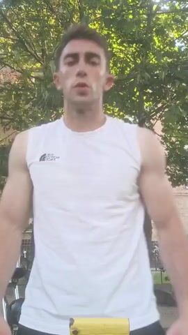 A young man with short brown hair and a light complexion is standing outside. He is wearing a white sleeveless t-shirt with a black logo on the left side of his chest and black shorts. He has a yellow object in his hands and is looking at the camera. The background shows a leafy green tree and a brick building. The sun is shining, casting shadows. The man appears to be in the middle of an exercise routine. His expression is focused, and he seems to be exerting himself. The setting appears to be a residential area, possibly a backyard or a park, given the presence of the tree and building. The overall mood is energetic and active. The logo on his shirt is partially visible but not clearly discernible. There is no other visible text in the frame.