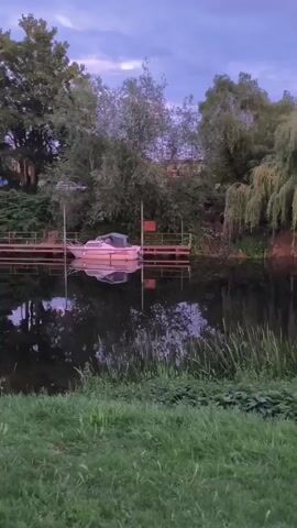 The video pans across a narrow canal bordered by green grass and reeds. Several narrowboats are moored along the canal. On the left, a boat is partially covered with white tarpaulin. Further down, a bright turquoise narrowboat is docked. On the right side of the canal, a vibrant pink narrowboat is visible. Beyond the canal, houses with red-tiled roofs and trees are situated. The sky is overcast with shades of purple and grey, suggesting it is dusk or dawn. The water of the canal reflects the sky and surrounding objects.