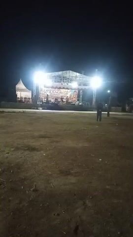 The video shows an outdoor concert stage at night. The stage is lit with bright lights, and there are speakers set up. A banner with text is visible on the stage. Several people are standing in front of the stage, and a few more are visible in the background near a tent. The ground in the foreground is dirt and grass.