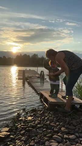 A woman and a young child are standing on a wooden dock over a body of water at sunset. The woman, wearing a striped tank top and blue pants, is leaning down and pointing at something in the child's hand. The child, wearing a t-shirt and green pants, is looking at their hand. The sun is setting over the water, casting a warm glow on the scene. There are trees and clouds visible in the background. The shore in front of the dock is covered in rocks and some grass.