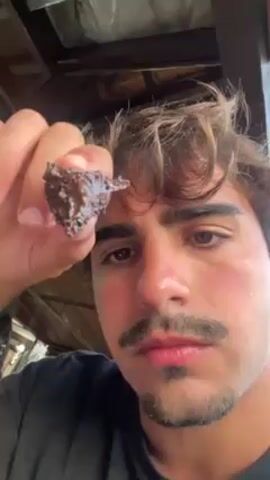 A young man with light brown hair, a mustache, and a goatee is the main focus of the video. He is holding a small, dark, irregularly shaped object, possibly a piece of food, in his right hand, close to the camera. He appears to be indoors, under a wooden roof structure. He brings the dark object to his mouth and eats it. He chews with a slightly displeased expression.