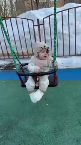 A baby, dressed in white and beige heart-patterned clothing and a matching bonnet, is on a swing at a playground. The swing has green rope supports and a red and black seat with a safety bar. A hand pushes the swing, and the baby laughs and smiles while being swung. There is snow piled up behind a metal fence in the background. The playground surface is green and blue.