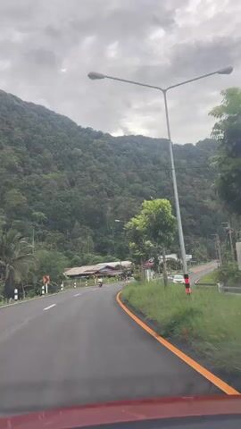 A car is driving along a paved road that curves to the right. The road is bordered by a grassy median with an orange stripe. On the left side of the road, there is a steep, tree-covered mountain. On the right side, there are some buildings and more trees. A person is riding a motorcycle in the same direction as the car. The sky is cloudy and overcast.