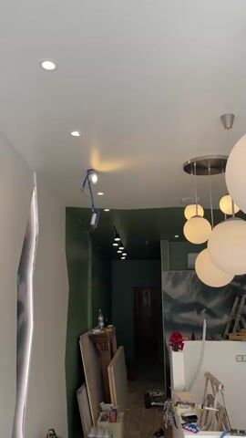 The video shows an interior space that appears to be under renovation. The ceiling is white with recessed lighting. A series of spherical pendant lights hang down from the ceiling, varying in size. The walls are painted a dark green in some areas and white in others. One wall features a large mural of a mountainous landscape. Construction materials, including boards and bottles of liquid, are visible on the floor. A ladder leans against a counter-like structure. The overall impression is of a commercial space undergoing a significant upgrade.