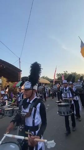 A marching band is parading down a street during the day. The band members are wearing black and white uniforms with tall black feathered hats. They are playing drums and cymbals. The street is lined with people watching the parade. The sky is clear and blue.