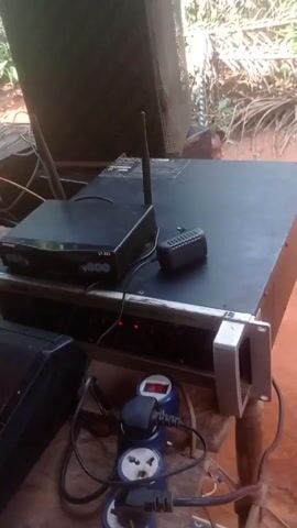 A close-up shot shows audio equipment set up outdoors. A black Shure UT-883 wireless receiver sits on top of a silver Power Amplifier P3300S. A black Yamaha keyboard is to the left. In front of the keyboard, a blue charging unit holds two blue batteries, a red light indicating they are charging. Multiple power cords are plugged into a blue power strip. Behind the equipment, a young child stands near a large speaker. The ground is red dirt. Foliage is visible in the background. Music is playing.