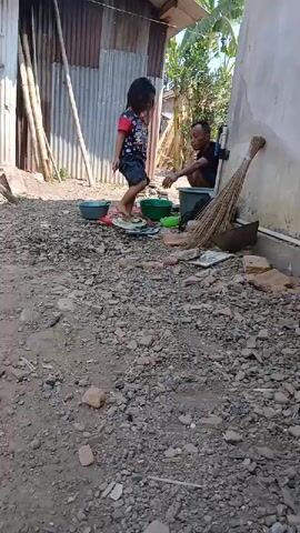 A young girl with dark hair, wearing a red shirt with a colorful print and a dark skirt, stands on a flat object on the ground. She is facing a man who is sitting and appears to be looking at something in a green bowl. The man is wearing a dark shirt. There are several plastic bowls on the ground near the girl, and a broom leans against a wall behind the man. The ground is covered with dirt, gravel, and small pieces of broken brick. In the background, there is a building with a corrugated metal wall and a wooden roof, and some tall bamboo poles. The scene appears to be outdoors during the day, with sunlight casting shadows.