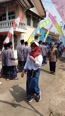 The video shows a group of young males, likely students, dressed in traditional Islamic attire. They are walking in a line on a dirt road, facing away from the camera. They are wearing grey outer shirts, dark pants, and black skullcaps. Some of them are also wearing sarongs. There is an older man in a patterned shirt and pants walking in front of them, also facing away from the camera. A woman in a red hijab and white shirt is walking towards the camera, also facing away from the camera. She is wearing a blue skirt and white sneakers, and has a black bag slung over her shoulder. The background shows a building with white trim and decorative flags strung across the street, suggesting a festive occasion. The lighting indicates it is daytime, with shadows on the ground.