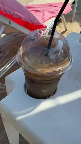 The video shows a close-up of a iced coffee drink in a clear plastic cup with a black straw. The cup is in a white plastic cup holder, which is part of a small white plastic table. In the background, there is a beach chair with a pink towel on it, and sand. The drink appears to be frothy and cold. The lighting suggests it is daytime and sunny. There are no people visible in the video.