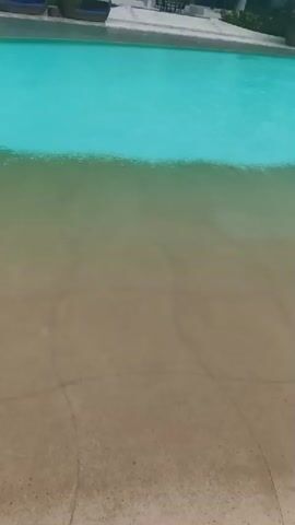 The video shows a shallow pool area with a sandy bottom. The water is a bright blue and transitions to a sandy color near the edge. In the background, there are lounge chairs and a building with balconies. A person is visible in the water near the edge of the pool, appearing to be swimming or wading. The scene suggests a resort or hotel pool area.