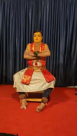 A man in elaborate Kathakali costume and makeup sits cross-legged on a red carpet in front of a black curtain. He has yellow face paint with red accents around his eyes and on his lips. He wears a red and gold vest, a white garment draped over his lower body, and black pants. His wrists and ankles are adorned with beaded bracelets. His arms are crossed in front of him, and he holds a green object in his hands. The video captures the man sitting in a static pose, with the accompanying audio featuring percussive musical accompaniment.