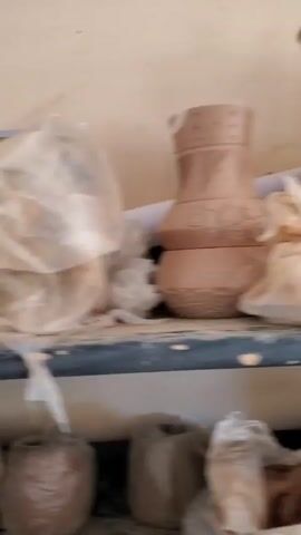 The camera pans across dusty shelves holding terracotta pots and vases in an indoor space. The shelves are constructed of dark metal supports. The pots are a light brown color and many of them feature carved or etched designs.  Some are stacked on top of each other.  Some items are covered in white plastic bags or dark plastic sheets.  A white board is visible in the background.  The room appears to be a pottery studio or workshop.