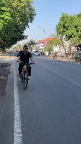 A man rides a bicycle down a paved road. He is wearing a dark cap, a dark short-sleeved shirt, dark pants, and sandals. He is pedaling the bicycle, which is yellow and black. The man is on the left side of the road, with a white line marking the edge of the lane.

The scene is outdoors, with trees lining the left side of the road and buildings on the right. The sky is clear and blue, suggesting a sunny day. The sun appears to be low in the sky, casting long shadows, indicating it is either early morning or late afternoon.

In the background, there are cars and motorcycles on the road. The buildings on the right are varied, with some appearing to be shops or businesses. One building has a red and white sign with the Honda logo.

The overall mood of the image is peaceful and ordinary, depicting a common scene of daily life. The location is identified as Gelam, Indonesia. There is no visible text in the image that can be clearly deciphered.