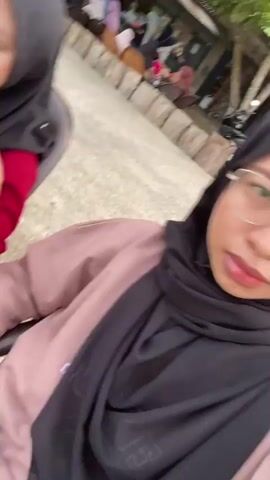 This media file captures a candid moment of two young women outdoors, likely during daylight hours, given the bright lighting and the presence of lush greenery. The primary subject, a young woman wearing a dark hijab and a bright red long-sleeved shirt, is seated in a dark chair. She is holding a translucent cup with a green liquid and a straw, appearing to be drinking from it. Her gaze is directed slightly away from the camera, and her expression is neutral.

The second individual, whose face is not fully visible, is also seated, wearing a muted pink sweatshirt. The camera appears to be held by this second person, suggesting a selfie-style recording or a shot taken by a companion.

The setting is an outdoor area with paved walkways and scattered tables and chairs, indicating a public space like a park, cafe, or recreational area. In the background, several other people are visible, some seated and others standing, further suggesting a gathering or a public venue. Trees and foliage frame the scene, contributing to the natural, possibly tropical, ambiance. The ground surface is concrete, and there are some wooden structures and benches visible.

The overall mood is relaxed and casual, typical of people enjoying a drink and spending time together outdoors. There is no visible text in the media file. The mention of "Air Molek, Indonesia" provides a specific geographical context for this scene.