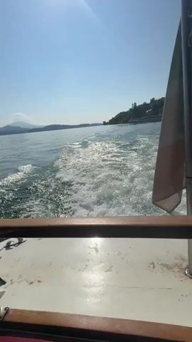 The video shows a sunny boat ride on a large body of water, likely a lake, near Binda, Italy. The initial shot, from the stern of the boat, captures the frothy white wake trailing behind. An Italian flag, green, white, and red, is prominently displayed on a pole at the back of the boat, fluttering gently in the breeze. In the background, a verdant hillside dotted with buildings stretches along the shore under a clear, bright blue sky. The sunlight creates a noticeable glare on the water. As the video progresses, the view shifts to the side of the boat, revealing vast expanses of calm blue water and distant, majestic mountains beneath the same cloudless sky. The boat features a dark canopy and a polished wooden railing. No people are visible, and no text is present in the video.