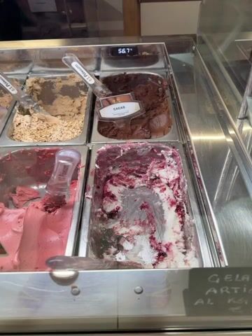 The camera pans slowly across a gelato display case with several flavors of gelato. The gelato flavors visible include a white flavor with a purple swirl, strawberry, Zuppa Inglese, pistachio, Gianduiotto, Cioccolato Fondente, mandorla, and cacao. Each flavor has a clear plastic serving spoon inserted into it, and the flavors are labeled with small signs. The temperature of the display case reads 56.7 degrees. The display case is well-lit and appears to be inside a gelato shop.