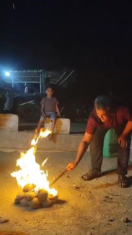 A group of people are gathered around a fire at night. The fire is burning brightly and there are several coconuts in the flames. A man is standing next to the fire, holding a stick and tending to the flames. A child is sitting on a ledge behind the fire, watching the flames. In the background, there are several motorcycles and a building with lights on. The scene appears to be outdoors, possibly in a village or rural area.