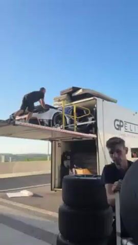 Here is a description of the video:

The video shows two men in front of a white GP Elite trailer. The man closest to the camera is leaning on a stack of tires and looks at the camera. The other man is working on a white race car loaded on a ramp that extends from the trailer's top level. The trailer has a logo that says “GP ELITE” and “DRIVE TO PERFECTION” on the side. The ramp is painted white with a yellow railing, and it appears to be a garage on top of the trailer. There is a guard rail visible on the bridge in the background and a clear blue sky.
