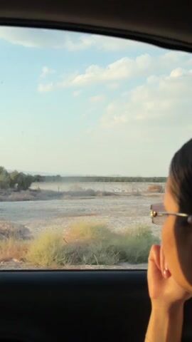 The video shows a view from inside a car, looking out the passenger side window. A person's face is visible in the foreground, turned away from the camera and looking out the window. The landscape outside appears to be arid, with sparse vegetation and dry brush. In the background, there is a sign with Hebrew writing on it, which is visible in the second frame. The sky is blue with scattered clouds, suggesting it is daytime. The car appears to be moving, as indicated by the slight blur of the background and the changing perspective. The overall impression is one of a journey through a dry, possibly rural or desert environment.