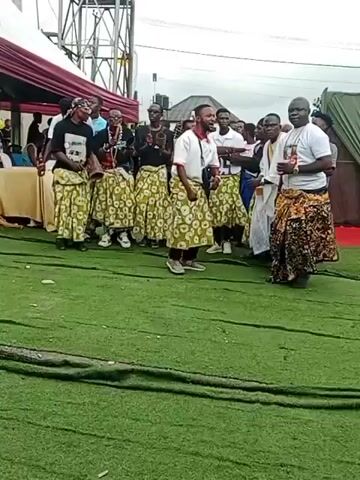 A group of people, some wearing matching green and white patterned skirts, are gathered outdoors on a green artificial turf. In the center, a man in a white shirt and green skirt faces the camera and appears to be singing or speaking. To his right, another man in a white robe and patterned skirt stands facing him. To the left of the central man, a group of individuals, some also wearing the green and white skirts, are clapping and looking towards the center. In the background, a large white tent structure with red trim is visible, along with a tall metal tower. The sky is overcast.