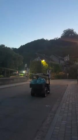 A golf cart carrying blue trash bags drives down a road next to a cobblestone sidewalk. The road appears to be a bridge crossing over a body of water. In the background, there are buildings on a hill covered in trees. The sky is clear and blue, indicating it is either early morning or late evening.