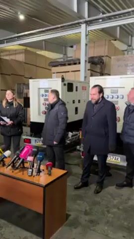 Two men stand in front of a table with many microphones, speaking to reporters who are not visible. Behind them are two electrical generators on trailers and stacks of cardboard boxes. The man on the left wears a black puffer coat, and the man on the right wears a dark overcoat. A woman stands to the left of the table, taking notes. The men appear to be giving a press conference about the generators. The room is a warehouse. The license plate on one of the trailers is visible, reading “WY 157AK”.