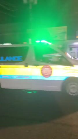 This video captures a scene at night, likely in Davao City, Philippines. The primary focus is a red tricycle with decorative stickers, including two stylized faces resembling the Joker. The tricycle is on a street, with other vehicles visible, including a bus with "STAR-03" written on its side and an ambulance with flashing green lights. Buildings and streetlights illuminate the scene, indicating it's nighttime. The overall atmosphere is one of a typical urban environment at night, with ongoing traffic. There are no discernible people in clear focus, but figures are present within the vehicles and on the sidewalks. The video is handheld and somewhat blurry, contributing to a dynamic feel.