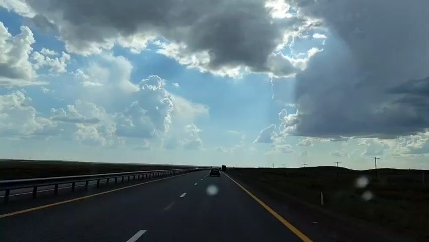 A car is driving down a highway. The sky is cloudy, with some sun peeking through. The landscape is flat, with some grass and trees. There are other cars on the road, but they are far away. The car is driving on the right side of the road. The road is paved and has white lines marking the lanes. There is a guardrail on the left side of the road.