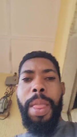 A man with a beard is looking at the camera. The background appears to be outdoors with a yellow wall on the left and a curtain on the right. In the background on the left, there is an electrical box mounted on the wall. The man is speaking and gesturing with his hands. The video seems to be a selfie video.