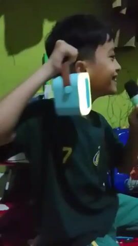 A young boy with dark hair wears a dark green t-shirt with a yellow number 7 on it and green pants with yellow stripes. He is sitting and holding a light blue speaker with a brown handle in his left hand, and a blue microphone in his right hand. He is smiling and appears to be singing or talking into the microphone. The speaker has a bright white light on one side. The background is a lime green wall with some shadows and what appears to be toys on shelves. A box with Iron Man on it is visible to the boy's right. The scene is dimly lit.